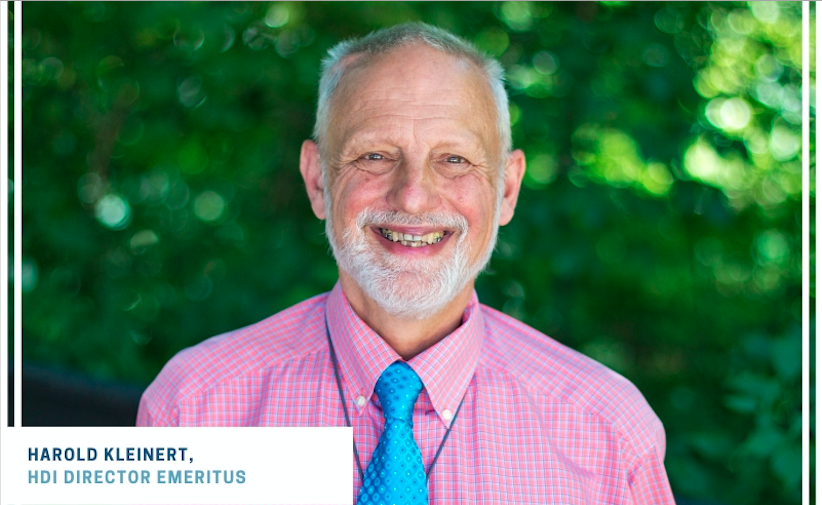 Harold Kleinert with white hair and beard smiling at the camera wearing a pink shirt and blue tie.