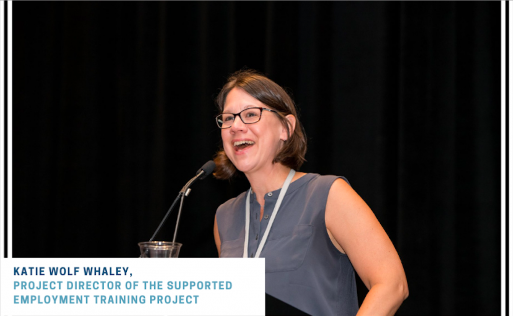 Katie Wolf Whaley Photo. she is wearing a grey dress and speaking using a microphone in front of a black background