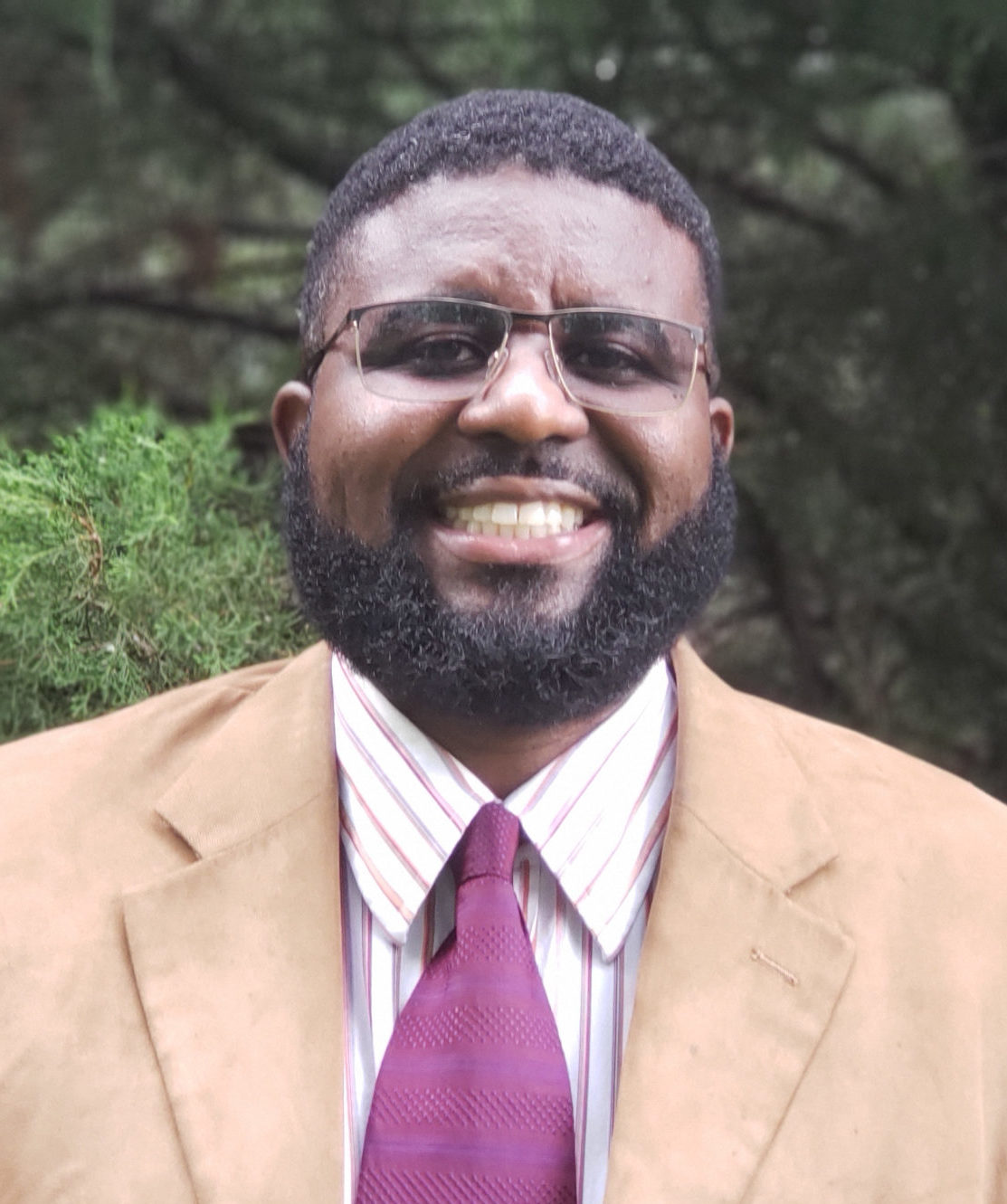 Lonnie is smiling at the camera in front of trees. He is wearing square-framed glasses, a khaki sportcoat, white striped dress shirt, and a maroon tie.