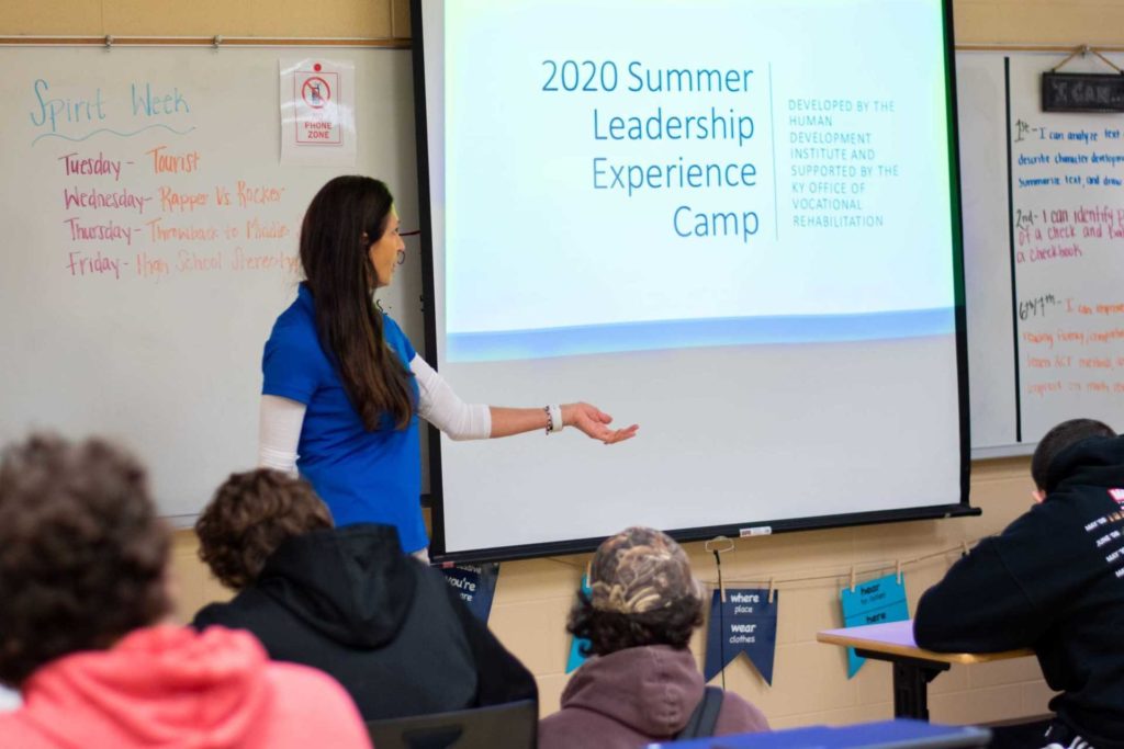 Woman with long brown hair wearing blue tshirt presenting to a class