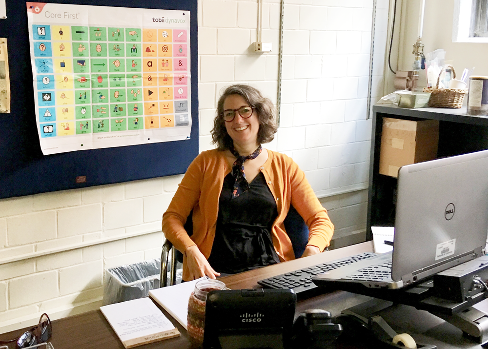 A woman with brown hair and black glasses wearing a black dress and copper sweater seated at a brown desk with white papers on it. A silver laptop is on her desk next to office supplies and a telephone. There is a multi-colored chart behind her.