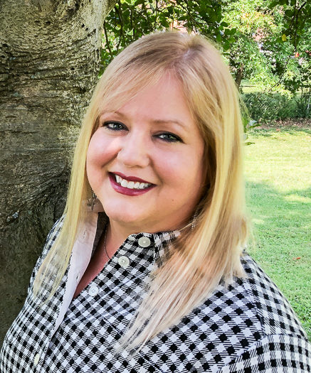 Kristen wearing a black and white gingham shirt smiling in front of a tree trunk. She has long, blonde hair and is wearing berry lipstick