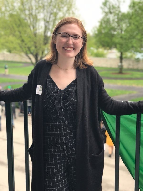 girl with red hair and black glasses wearing black dress and sweater standing in front of black railing with green trees and grass in the background