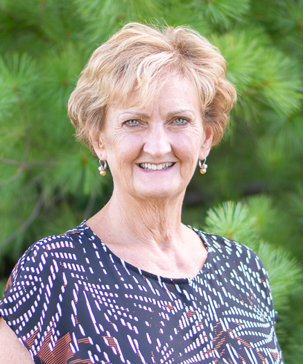 Shirley wearing a black, white, and red shirt smiling at the camera in front of trees