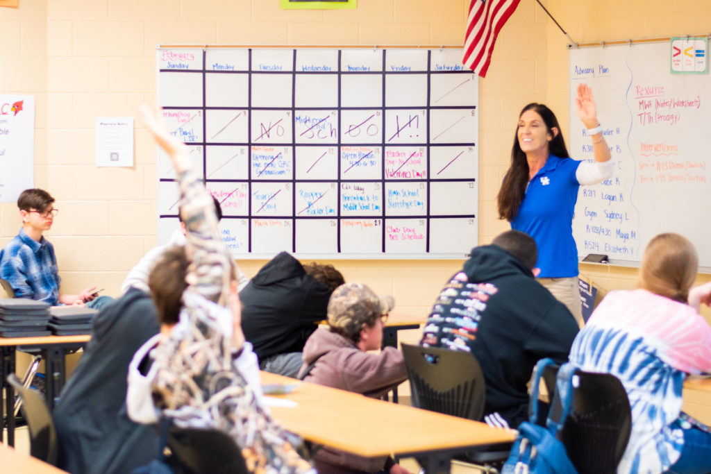 Woman with long brown hair wearing a blue shirt in front of a classroom of 7 students wearing a black sweatshirt, burgundy sweatshirt and hat, black sweatshirt with multi-colored lettering, tie dye blue and pink shirt, blue flannel shirt, navy sweatshirt. One student, wearing a white sweatshirt with black cartoons on it, has his hand raised.