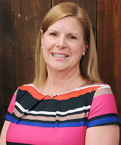 Kimberly standing in front of a wooden wall smiling at the camera. She has shoulder-length hair and is wearing a colorful, striped shirt.