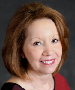 Dr. Capilouto is smiling on front of a grey background. She has shoulder-length, red hair and is wearing red lipstick.