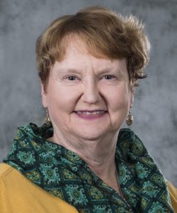 Jane Kleinert headshot. She is wearing a yellow sweater with a green, paisley scarf. She has short, red hair. She is also wearing gold earrings.