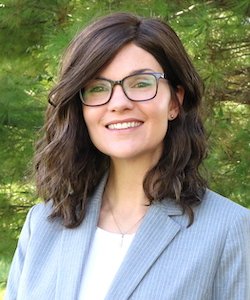 Jessica Ware staff photo. She has shoulder-length curly brown hair and is wearing glasses, a white blouse, and grey blazer