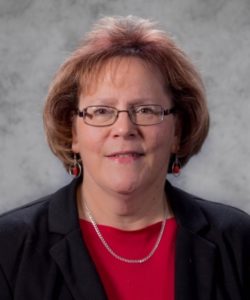 Judith Page headshot. She is wearing a red shirt and black blazer with a silver necklace and red bead earrings. She has short, auburn hair. She is wearing glasses.