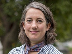 Emily Moseley, a white woman is wearing a blue and white striped shirt and scarf. She has dark, shoulder length brown hair and is smiling. There are trees in the background.