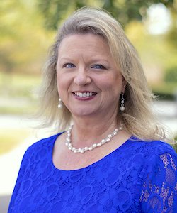 Alison Heitmeyer staff photo. She is wearing a UK blue blouse with pearl necklace and earrings. She has long, blonde hair, standing in front of trees, and smiling at the camera.