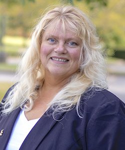 Elizabeth Thompson staff photo. She has long, curly blonde hair and is wearing a white shirt and black blazer. She is standing in front of trees and smiling at the camera