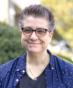 Erin Fitzgerald staff photo. She has short, grey hair, is wearing a blue button down and necklace. They are standing in front of trees and smiling at the camera.