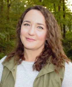 Hannah Hyatt staff photo. She is wearing an ivory blouse with a green vest and standing in front of trees. She has shoulder-length curly, brown hair and is smiling at the camera