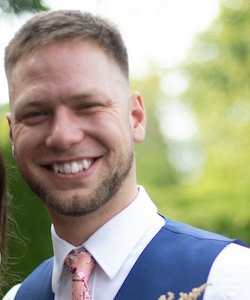 Jon Watkins staff photo. He is wearing a white collared shirt with a blue vest and a pink floral tie. He has shirt, brown hair and a beard. He is smiling.