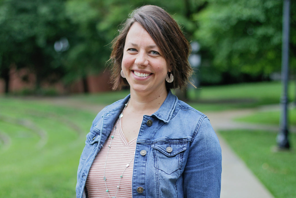 smiling woman with short brown hair wearing a tan top, jean jacket, and hoop earrings with trees in the background