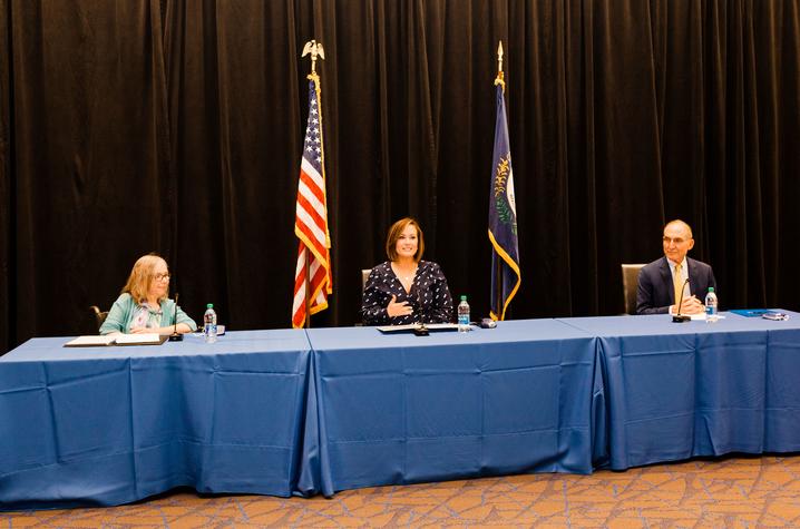 two women and one man seated at a table with blue tablecloths on it. They are seated in front of the American and Kentucky flags. The woman on the left has blonde hair, is wearing a light blue top and sweater. The 2nd woman is wearing a navy blue dress; and the man is wearing a navy blue jacket and a yellow tie.