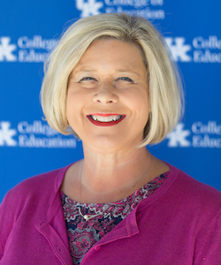 Jennifer Grisham staff photo. She is wearing a pink paisley blouse with a pink sweater and is standing in front of a UK backdrop. She has short, blond hair and is smiling at the camera.