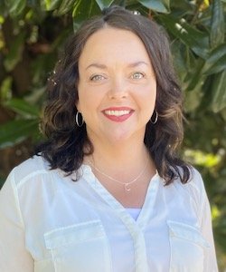 Kristy Ryan-Goodwin Staff photo. She is wearing a white blouse and has shoulder-length curly hair. She is standing in front of a tree and is smiling at the camera.