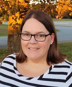 Amy Drake staff photo. She is wearing a black and white striped shirt and black-rimmed glasses. She has shoulder-length brown hair