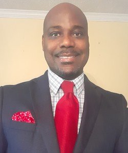 Anthony Glenn wearing a grey suit with a red tie and hanker-chief. He is smiling at the camera