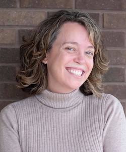 Candace Storrer staff photo. She is wearing a beige turtleneck sweater and standing in front of a brick wall. She has long, wavy light brown hair and is smiling at the camera