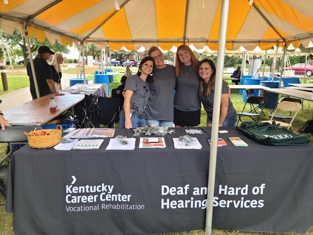 4 women standing behind a conference table under a tent