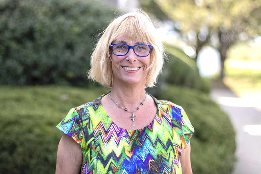 Tina Lindon staff photo. She is wearing a colorful chevron blouse and necklace. She is standing in front of trees and smiling at the camera.