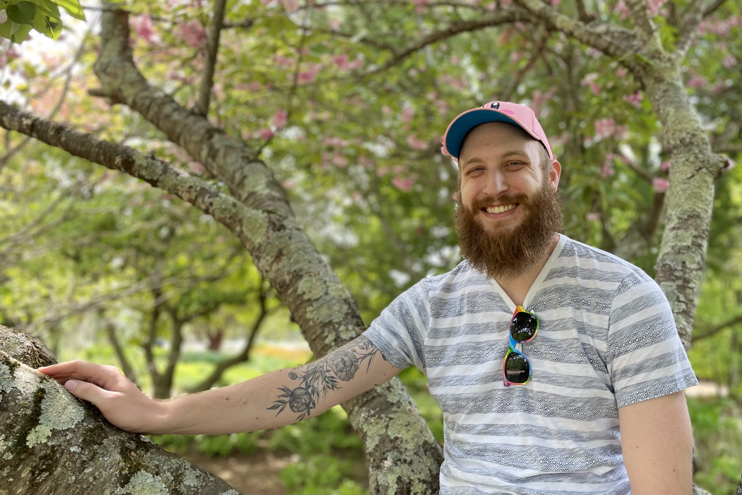 man wearing a striped shirt, sunglasses, and a hat standing in front of trees