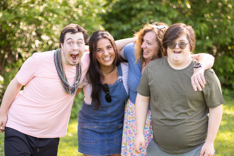 Four people smiling and laughing with their arms around each other. Trees are in the background.