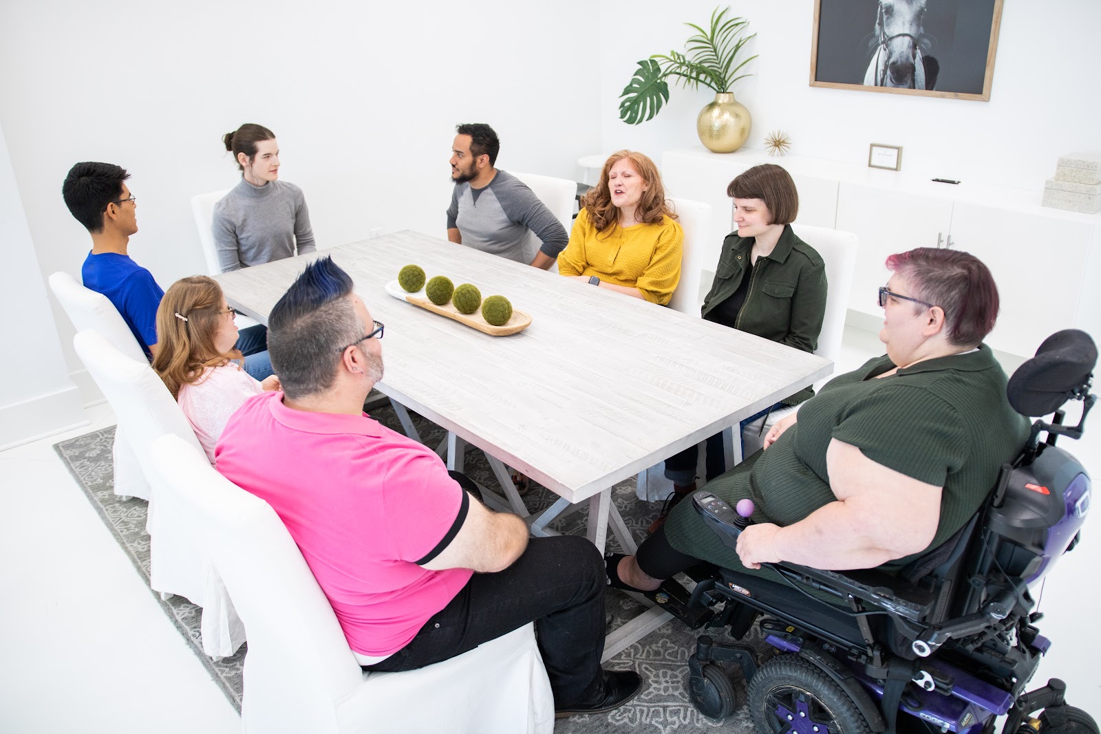 A group of people sit at a table in a bright office space. A few members of the group are in conversation while the others listen. 