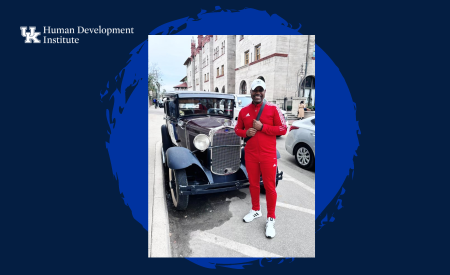 Anthony Glenn wearing red pants, red shirt, and a white hat standing in front of an old black car on a street